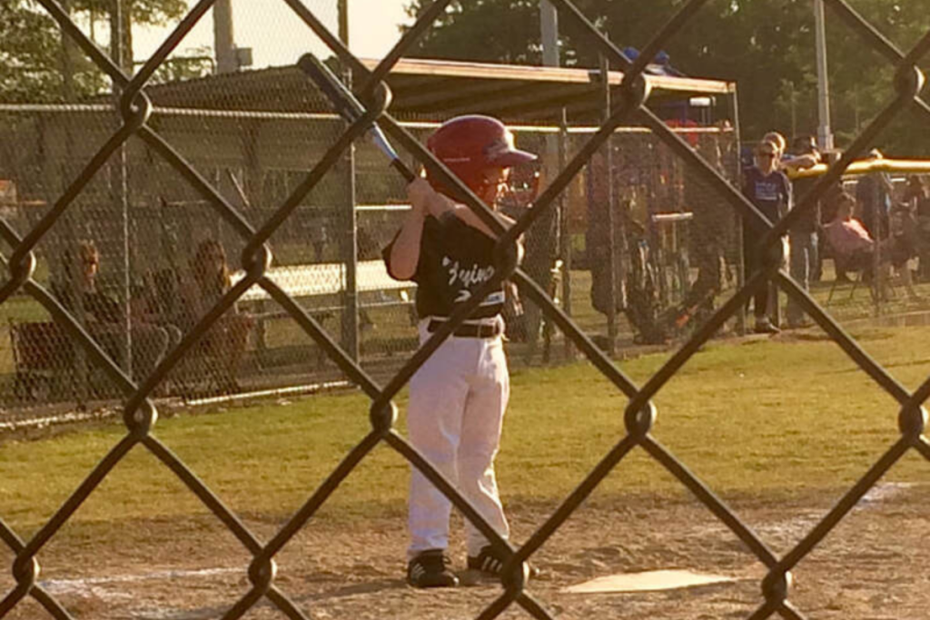 A boy stands at the baseball mound ready to swing, while his mother - who suffers from Anxiety - watches from behind a chain-link fence.