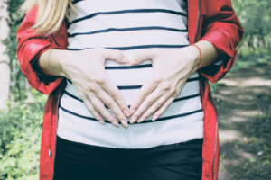 A woman in a striped shirt and red coat creates a heart with her hands over a pregnant body, which was chosen to accompany the respective letter written to endometriosis at Dear Diagnosis: a literary project.